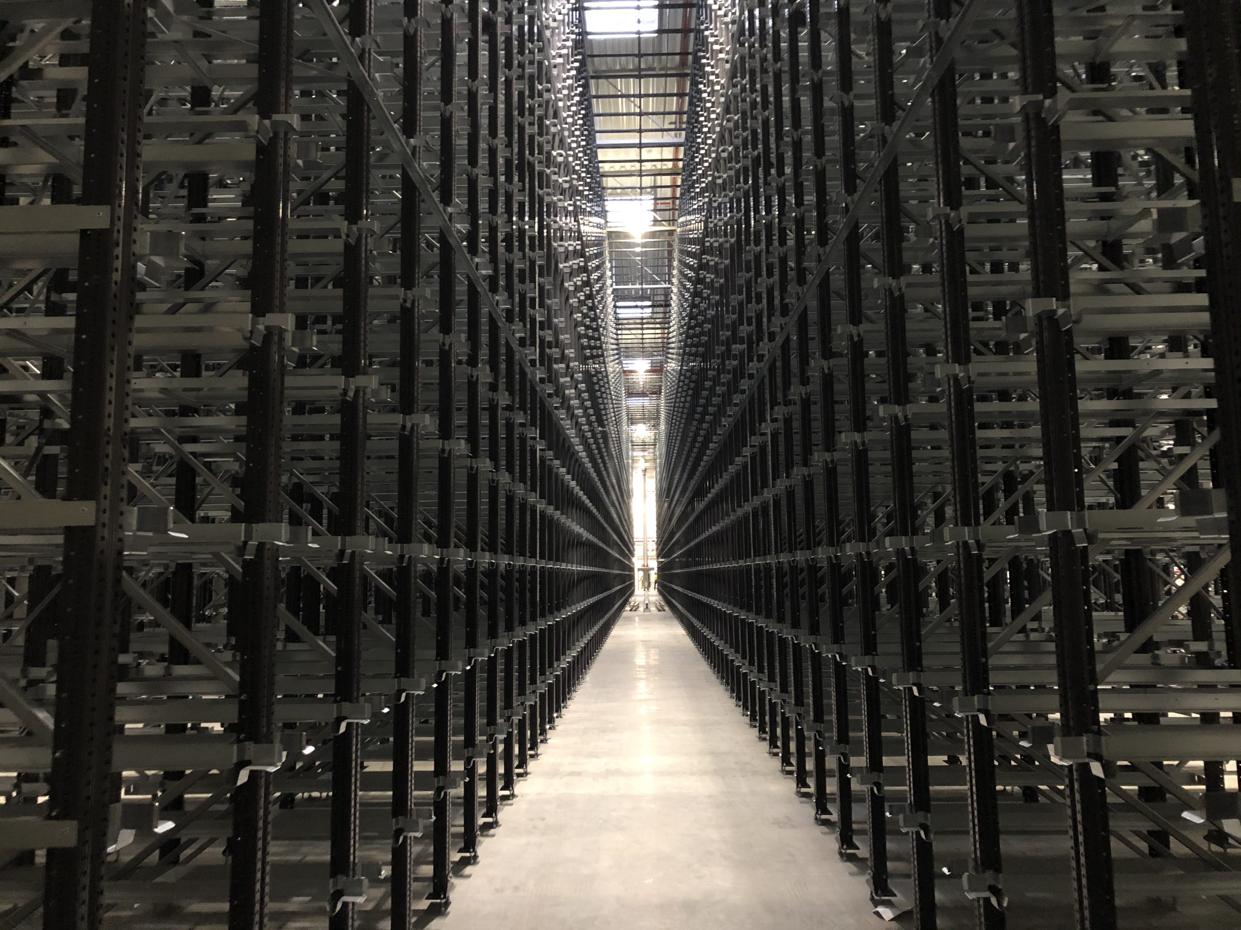 Wnętrze hali magazynowej z zamontowanymi, metalowymi regałami wysokiego składowania, widok w perspektywie z oświetleniem w tle.