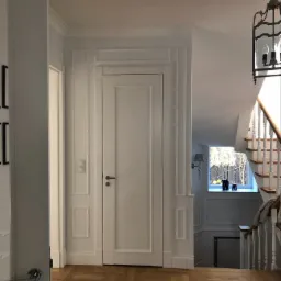 Wnętrze domu z parkietem w jodełkę, białe drzwi z dekoracyjnymi listwami, schody z białymi balustradami i widokiem na okno z figurkami słoni, wisząca lampa w stylu retro.