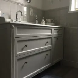 Szara szafka łazienkowa z trzema szufladami i umywalką, chromowana bateria, płytki w tle, okrągłe lustro na ścianie, elektryczna szczoteczka do zębów na blacie.