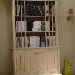 Kremowa, drewniana biblioteczka z przeszklonymi półkami wypełnionymi książkami i dokumentami, stojąca obok zielonego wazonu z pomarańczowym paskiem w pomieszczeniu o stonowanych ścianach.