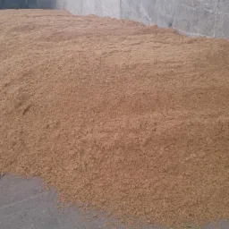 Góra jasnobrązowego, sypkiego materiału (prawdopodobnie zboża lub nawozu) na betonowej posadzce, z fragmentem opony ciężarówki w tle.