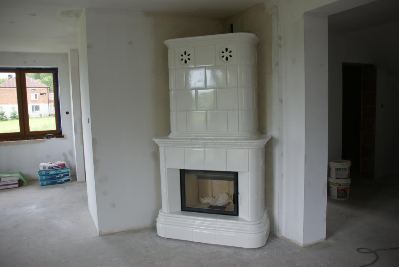 Biały, kaflowy piec kominkowy z czarnym wkładem, w surowym wnętrzu, widoczne worki z zaprawą i niedokończone ściany.