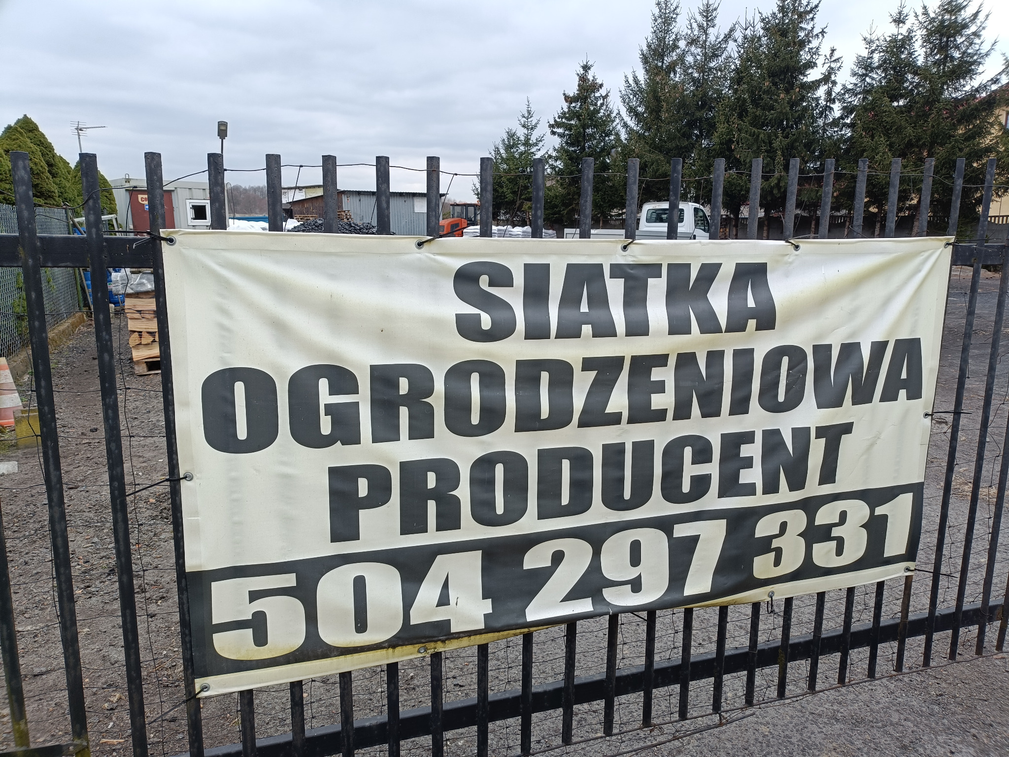 Baner reklamowy producenta siatki ogrodzeniowej z numerem telefonu, zawieszony na czarnym metalowym ogrodzeniu, na tle drzew i budynków.