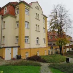 Widok z zewnątrz na odnowiony budynek z żółtą elewacją, czerwonym dachem i widocznymi śladami wilgoci w dolnej części ściany, sugerujący potrzebę osuszenia.