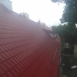 Czerwony dach z blachy trapezowej na budynku mieszkalnym, widok z góry, w tle drzewa i zaparkowane samochody.