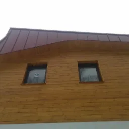 Dom z drewnianą elewacją i ciemnoczerwonym dachem z blachy, widoczne dwa okna z naklejkami na szybach.