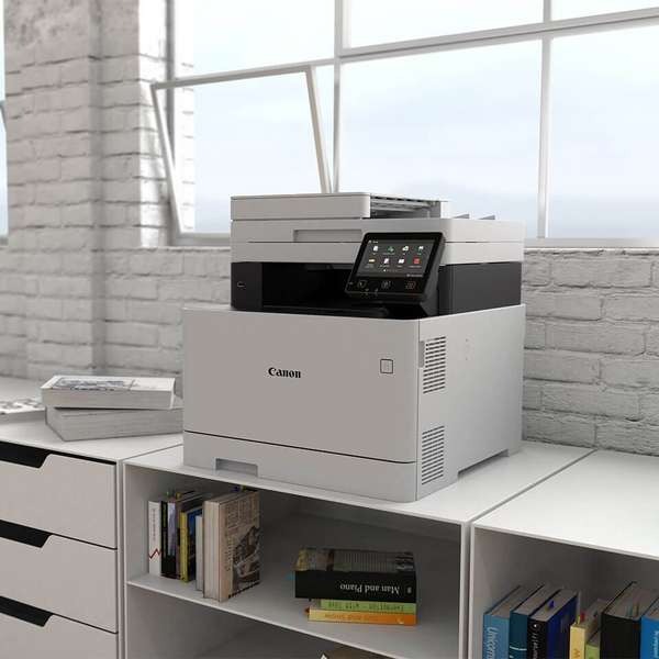 Nowoczesna, wielofunkcyjna kserokopiarka Canon na białej szafce, obok stos książek, widok z boku na tle ceglanej ściany i okna.