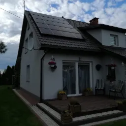 Szary dom jednorodzinny z brązowym dachem i panelami słonecznymi; widoczny numer domu '45', antena satelitarna i dekoracje kwiatowe.