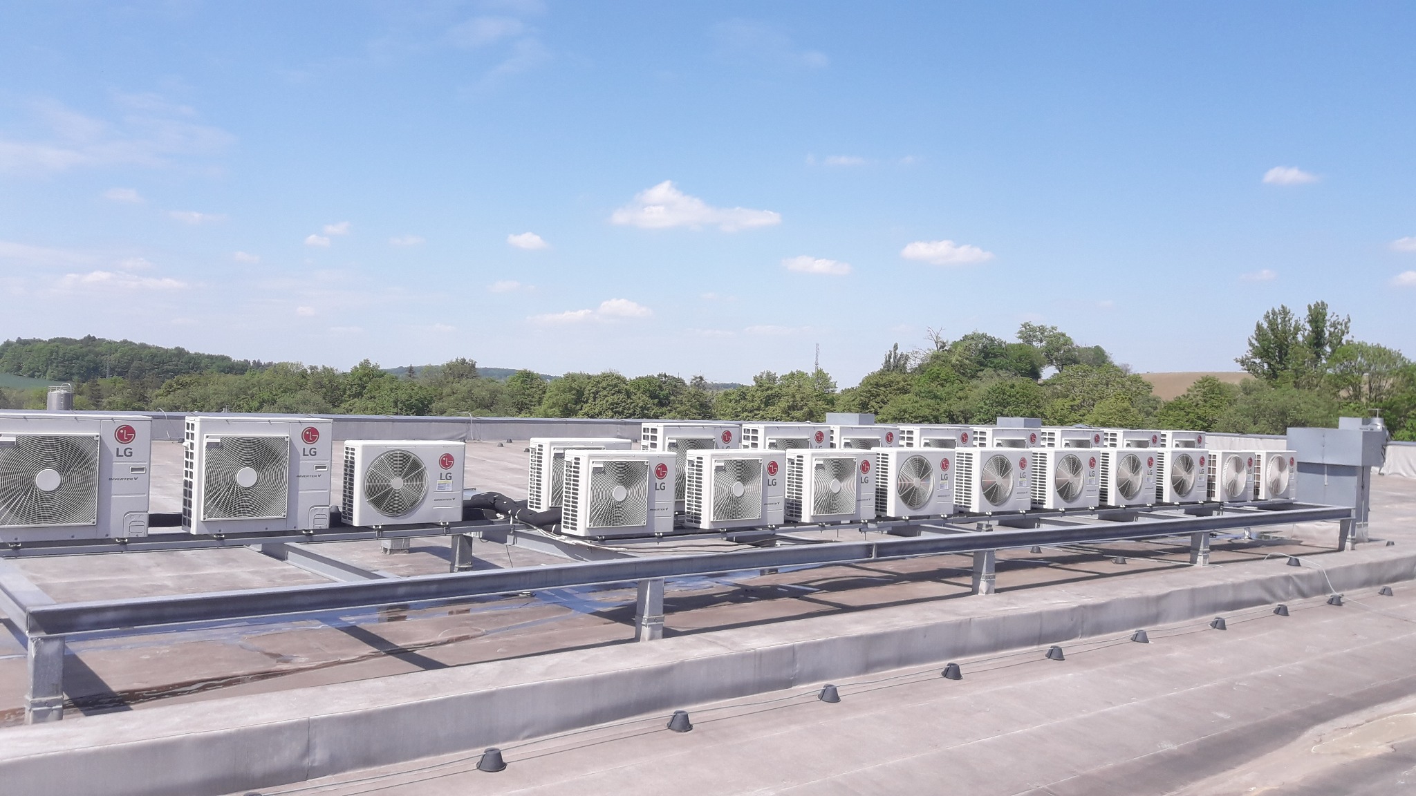 Rząd jednostek zewnętrznych klimatyzacji LG Inverter V, zainstalowanych na dachu budynku w Pogórzu, z widokiem na zalesione wzgórza w tle.