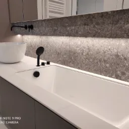 Nowoczesna łazienka z białą, prostokątną wanną i umywalką nablatową, czarna armatura, szare płytki na ścianie z podświetleniem LED, minimalistyczny design.