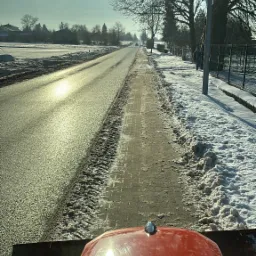 Widok z kabiny traktora odśnieżającego chodnik wzdłuż drogi w słoneczny zimowy dzień, z widocznymi śladami opon na odśnieżonej powierzchni i zaspami śniegu po bokach.