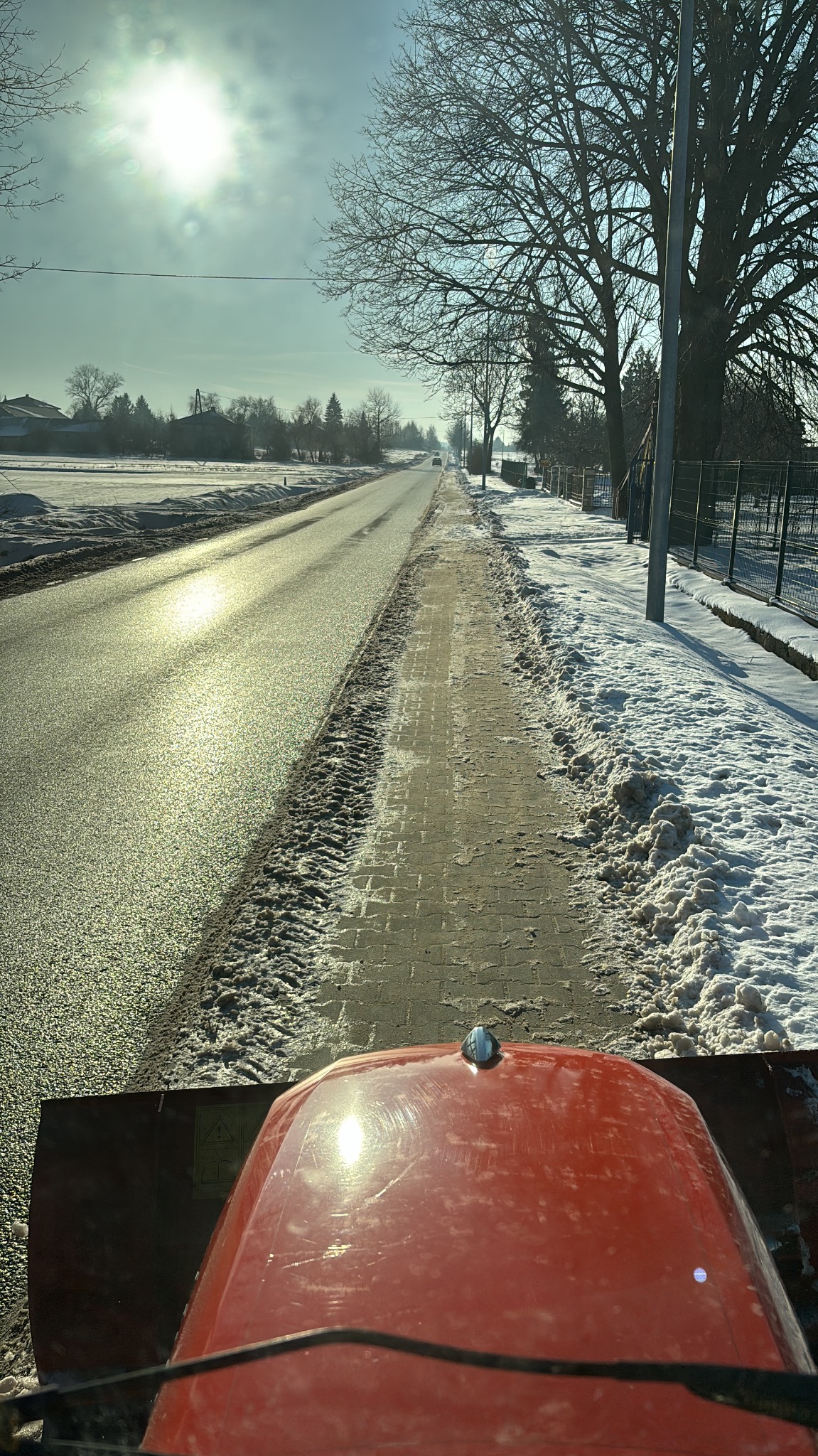 Widok z kabiny traktora odśnieżającego chodnik wzdłuż drogi w słoneczny zimowy dzień, z widocznymi śladami opon na odśnieżonej powierzchni i zaspami śniegu po bokach.