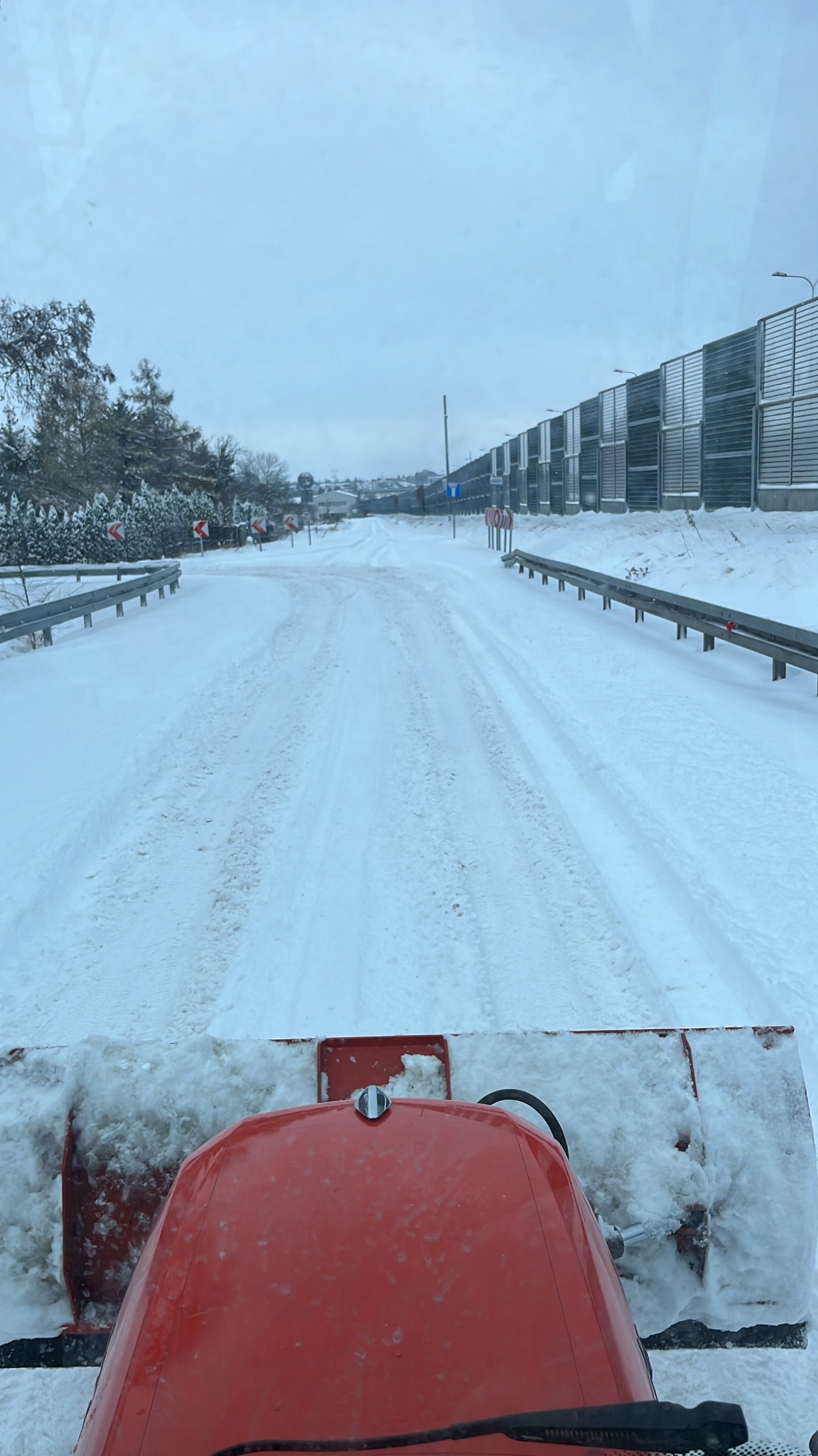 Widok z kabiny traktora na odśnieżoną drogę, z boku bariera dźwiękochłonna i drzewa pokryte śniegiem, pochmurny dzień.
