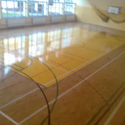Parkiet na sali gimnastycznej z wyraźnymi liniami boiska, odbijający światło z okien. Widoczny kosz do koszykówki i siatka ochronna.
