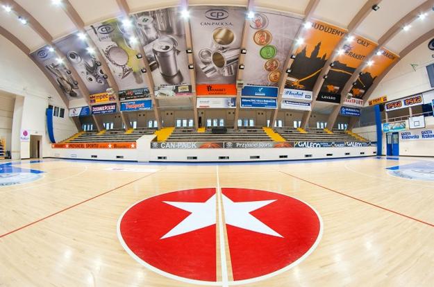 Jasne drewniane podłoże hali sportowej z czerwonym logo gwiazdy na środku. Trybuny w tle pod reklamami. Widok z perspektywy parkietu.
