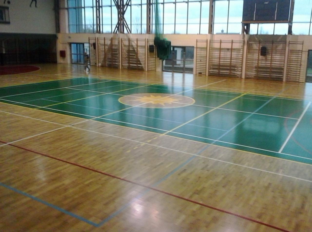 Sala gimnastyczna z parkietem w odcieniach brązu i zieleni, widoczne linie boiska i centralny wzór słoneczny. Duże okna i drabinki gimnastyczne w tle.