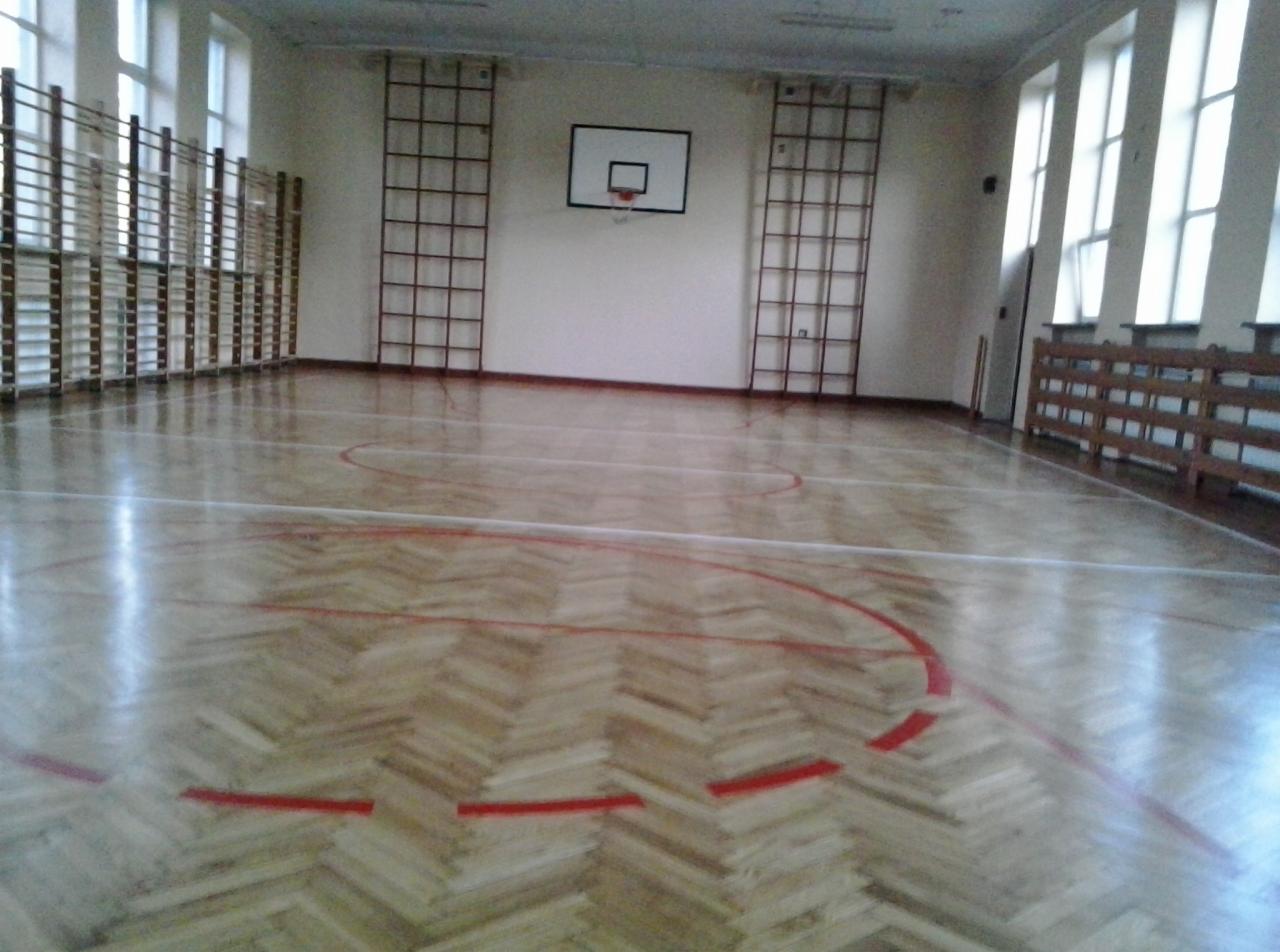Parkiet w jodełkę na sali gimnastycznej, z widocznymi liniami boiska, koszem do koszykówki i drabinkami gimnastycznymi przy ścianach, oświetlony przez okna.