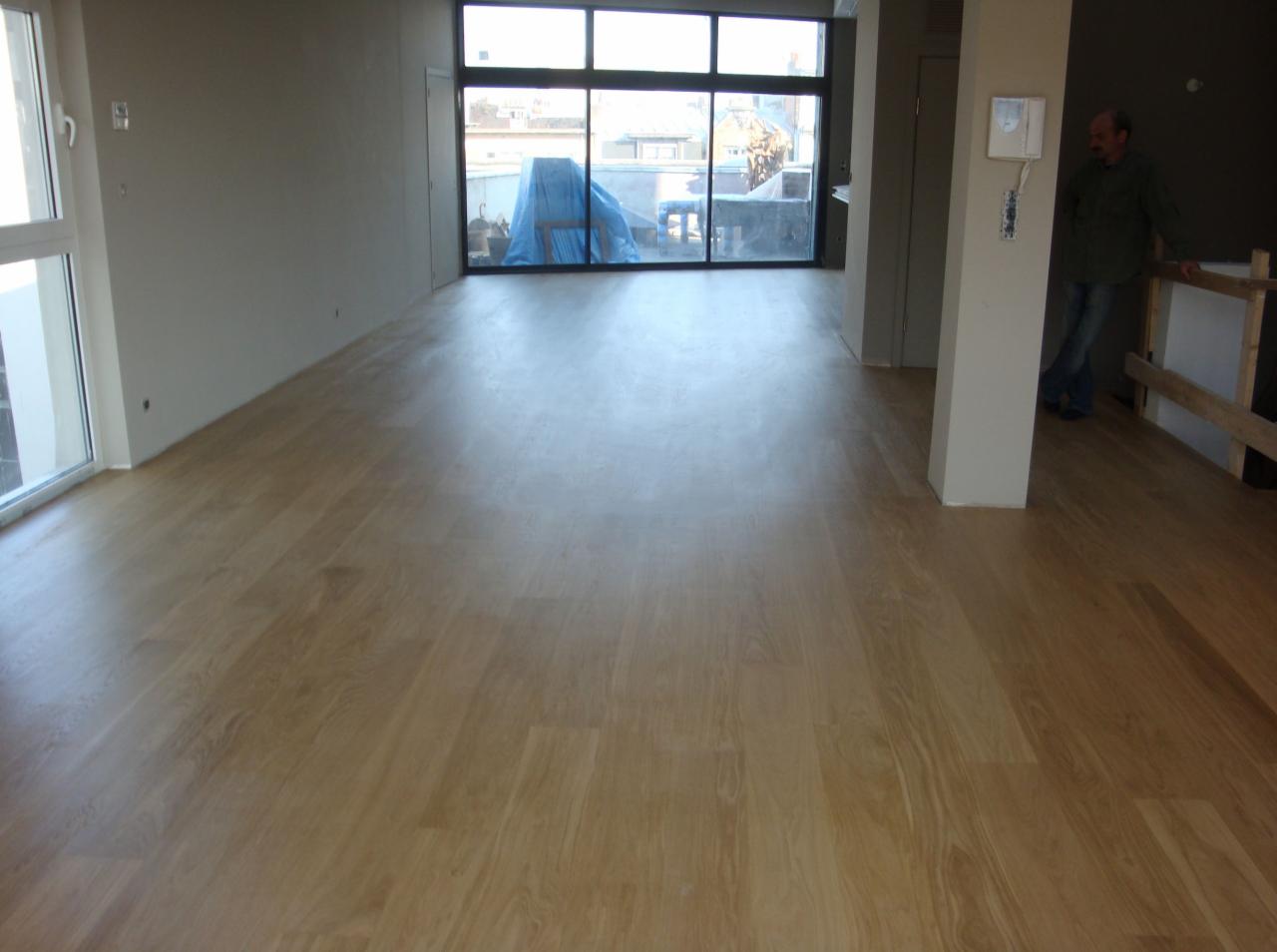 Jasny parkiet w przestronnym pokoju z dużym oknem balkonowym. Widoczny fragment remontowanego balkonu i osoba oparta o barierkę. Gładka powierzchnia podłogi.