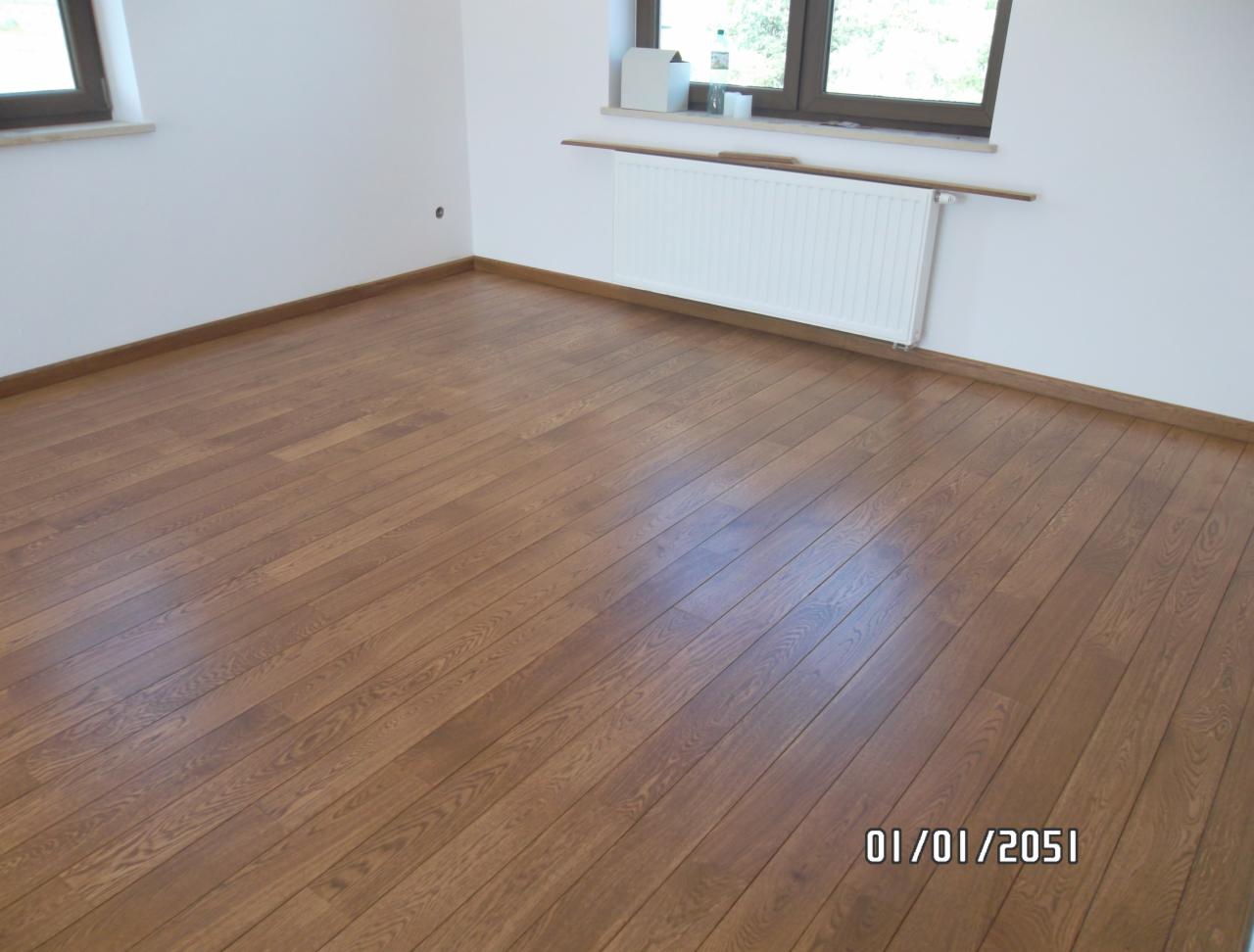 Salon z parkietem w odcieniach brązu, ułożonym w regularny wzór. Widoczne białe ściany, okno i grzejnik. Listwy przypodłogowe w kolorze drewna.