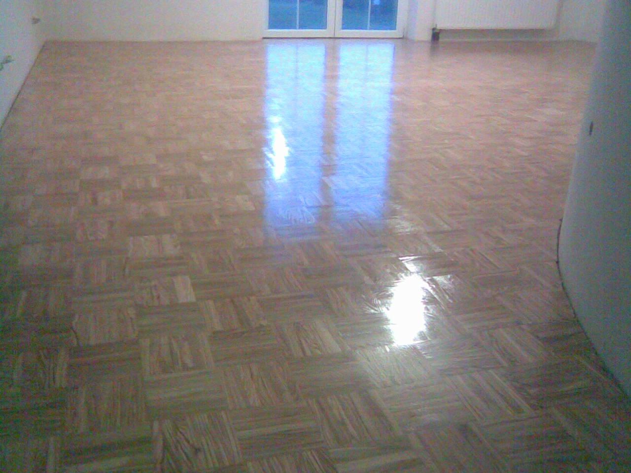 Ukończona podłoga z parkietu w jodełkę, świeżo po lakierowaniu. Widoczne odbicie światła od okna. Jasne wnętrze z białymi ścianami i grzejnikiem.