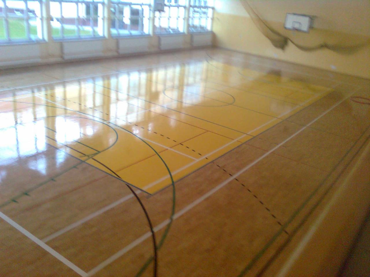 Parkiet na sali gimnastycznej z wyraźnymi liniami boiska, odbijający światło z okien. Widoczny kosz do koszykówki i siatka ochronna.