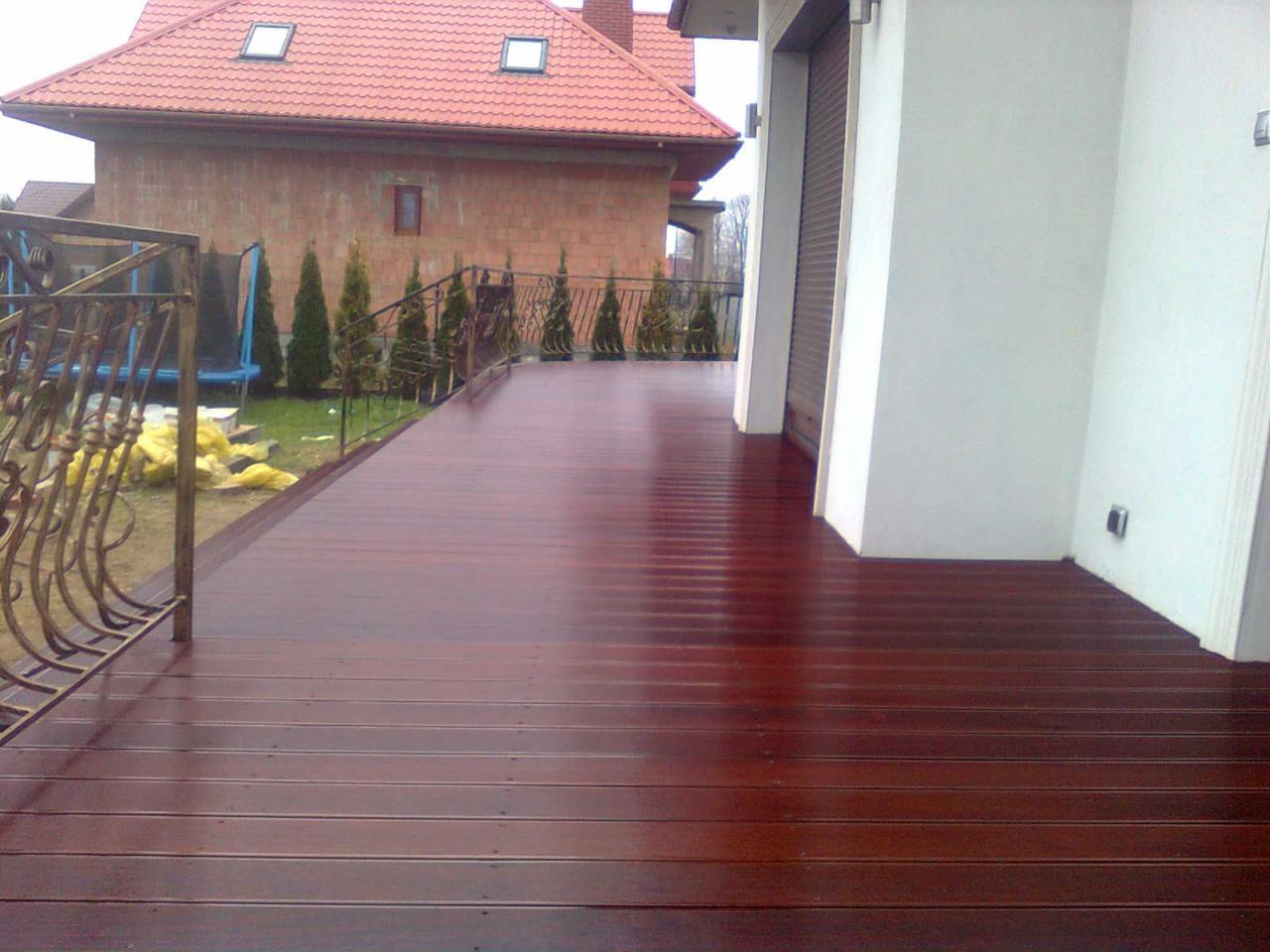 Błyszczący, brązowy taras z desek kompozytowych na tle domu z czerwonej cegły i zieleni. Metalowa balustrada z ozdobnymi detalami dodaje elegancji.