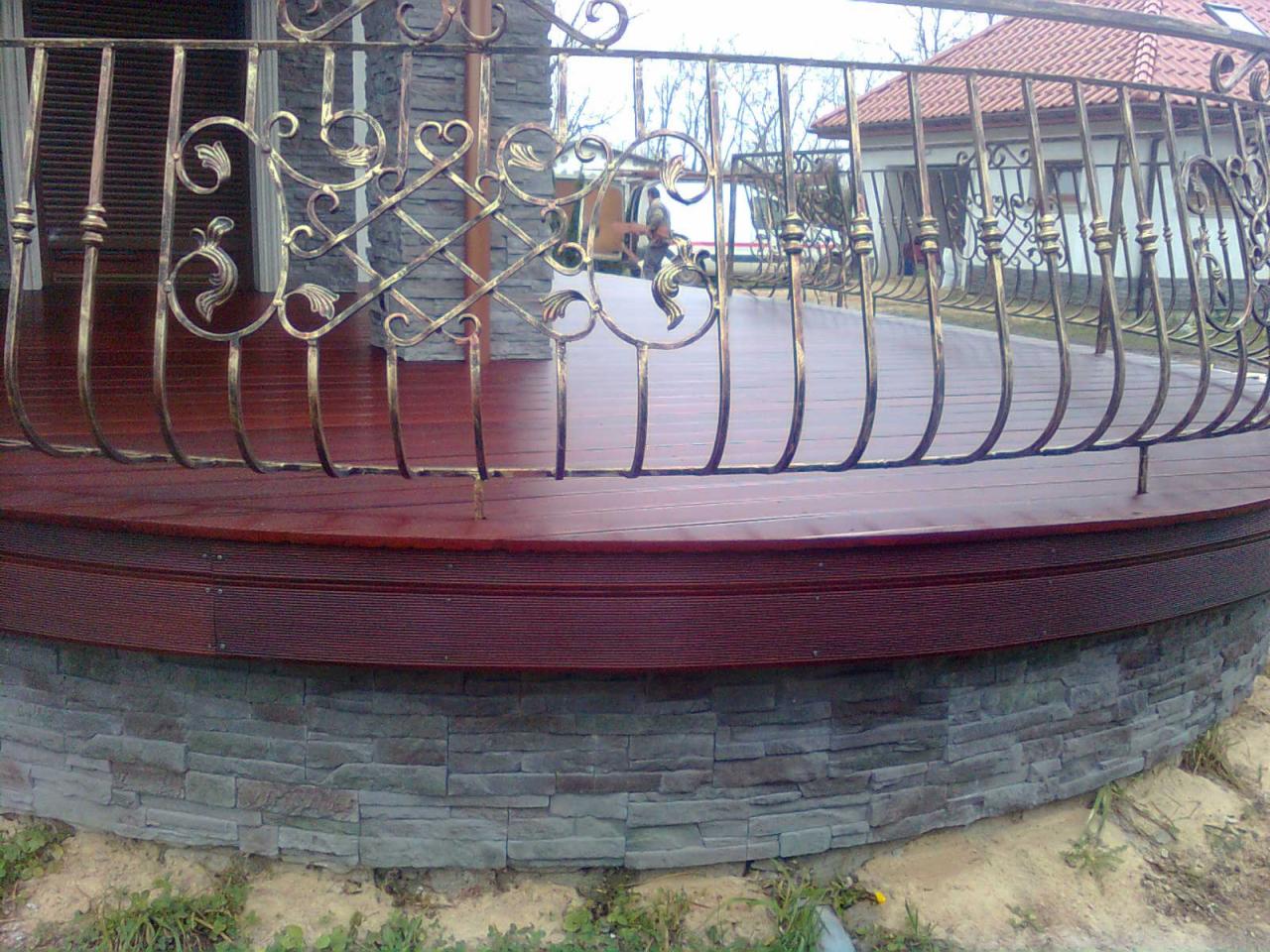 Drewniany taras z dekoracyjną, metalową balustradą i kamienną podmurówką. Widoczne elementy architektury ogrodowej i fragment domu w tle.