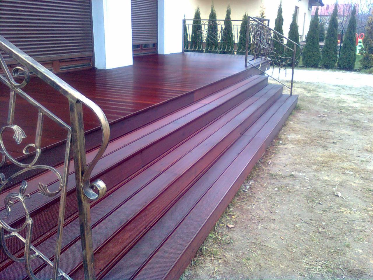 Ciemne, lakierowane drewniane schody zewnętrzne z metalową balustradą, prowadzące na taras. W tle widoczne tuje i budynek z roletami.