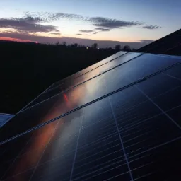 Panel fotowoltaiczny na dachu domu, widoczny na tle zachodzącego słońca i ciemnego pola.