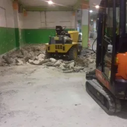 Wnętrze budynku w trakcie prac ziemnych: pomarańczowa mini koparka Kubota z widocznym numerem 140005 i żółta wywrotka na tle ściany z zieloną lamperią i gruzem.