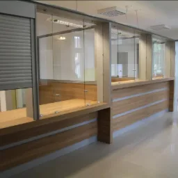 Wykonana na zamówienie lada recepcyjna z drewna i szkła, z roletą antywłamaniową, w jasnym, nowoczesnym wnętrzu. Widoczne detale mocowania szkła i poziome listwy dekoracyjne.