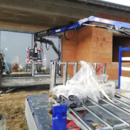 Montaż dużego okna za pomocą przyssawki i dźwigu na placu budowy, widoczny pracownik nadzorujący proces, surowy stan budynku w tle.