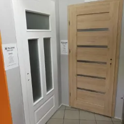 Dwie ekspozycyjne skrzydła drzwiowe wewnętrzne: białe z przeszkleniami i drewnopodobne z poziomymi pasami szkła, widoczne etykiety producenta.