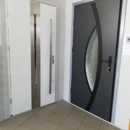 Ekspozycja dwóch modeli drzwi zewnętrznych: białe z pionowymi, metalowymi wstawkami i szare z owalnym przeszkleniem, prezentowane na tle kafelkowanej podłogi.