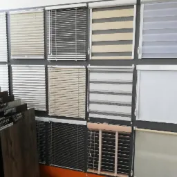 Ekspozycja różnych typów rolet okiennych, w tym plisowanych, żaluzji poziomych i rolet zwijanych, z widocznymi próbkami paneli podłogowych z lewej strony.