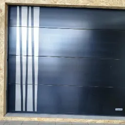 Czarna segmentowa brama garażowa z czterema poziomymi panelami i pionowymi srebrnymi paskami po lewej stronie, prezentowana wewnątrz pomieszczenia z widoczną drewnianą ramą i logo producenta...