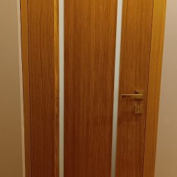 P.P.H.U. STOLAR SYSTEM ŁUKASZ ZIÓŁKOWSKI - Dębowa, fornirowana ościeżnica z prostokątnym przeszkleniem i złotą klamką. Widoczna faktura drewna i podłoga w podobnym odcieniu. Minimalistyczny design.