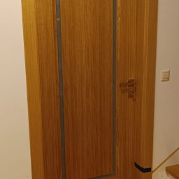 P.P.H.U. STOLAR SYSTEM ŁUKASZ ZIÓŁKOWSKI - Dębowa, fornirowana ościeżnica z szarą intarsją i klamką na tle białej ściany i drewnianej podłogi. Widoczne schody. Ciepłe oświetlenie.