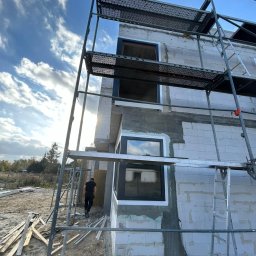 P.P.H.U. STOLAR SYSTEM ŁUKASZ ZIÓŁKOWSKI - Montaż okien w surowym budynku z białego bloczka, widoczne rusztowania i drabina. Czarna rama okienna kontrastuje z jasnym murem. Osoba w tle. Słoneczny dzień.
