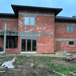 P.P.H.U. STOLAR SYSTEM ŁUKASZ ZIÓŁKOWSKI - Surowy stan budynku z czerwonej cegły z zamontowanymi oknami o drewnianych ramach. Widoczne niedokończone elementy konstrukcyjne i materiały budowlane na zewnątrz.