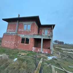 P.P.H.U. STOLAR SYSTEM ŁUKASZ ZIÓŁKOWSKI - Surowy stan dwukondygnacyjnego domu z czerwonej cegły z zamontowanymi oknami z brązowymi ramami, na tle pochmurnego nieba i zarośniętej trawy.