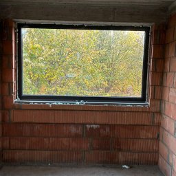 P.P.H.U. STOLAR SYSTEM ŁUKASZ ZIÓŁKOWSKI - Ciemna rama okienna w surowym murze z widokiem na jesienne drzewa. Montaż okna w trakcie budowy, widoczne pianki montażowe.