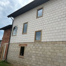 P.P.H.U. STOLAR SYSTEM ŁUKASZ ZIÓŁKOWSKI - Montaż okien w nowym domu z elewacją z białej cegły i ceglanym dołem. Widoczne ramy okienne z pianką montażową. Częściowo widoczna osoba i betoniarka.