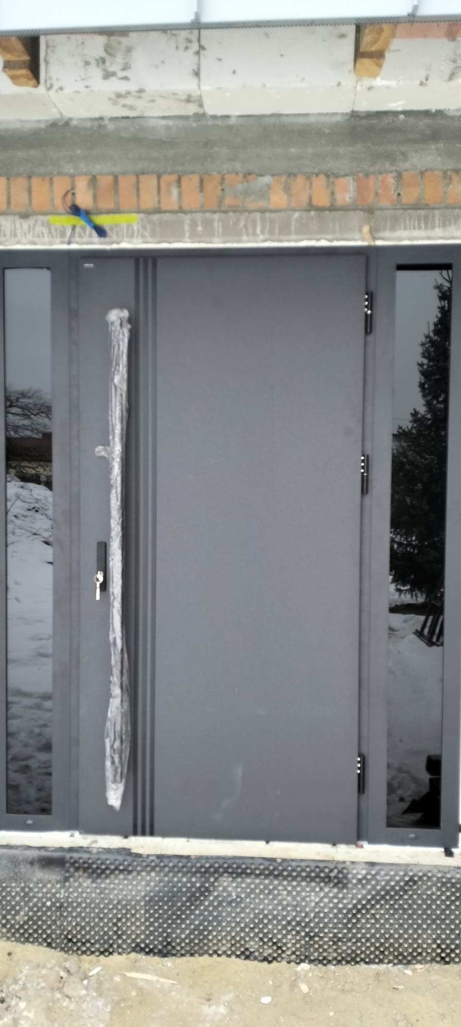 Szare drzwi wejściowe z bocznymi przeszkleniami, zamontowane w surowym stanie budynku. Klamka zabezpieczona folią. Widoczne elementy konstrukcyjne nadproża.