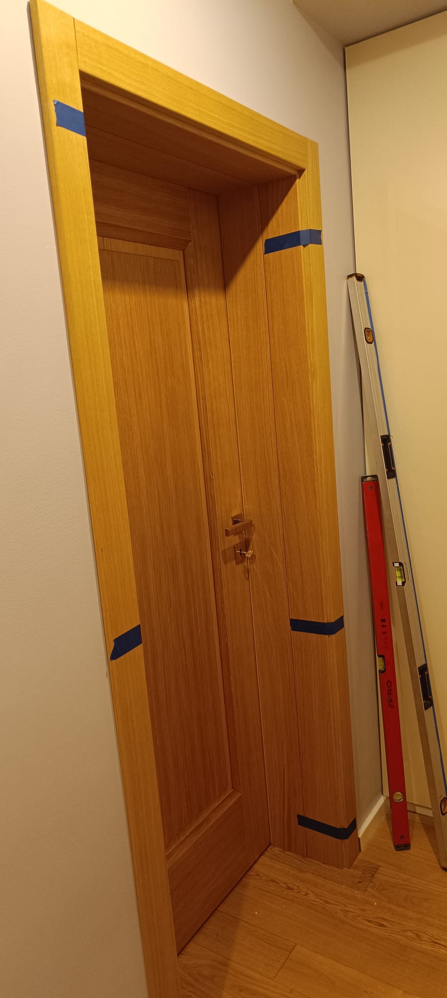 Zamknięte, drewniane drzwi wewnętrzne z niebieską taśmą zabezpieczającą ościeżnicę, wsparte o poziomice, po montażu w jasnym wnętrzu.