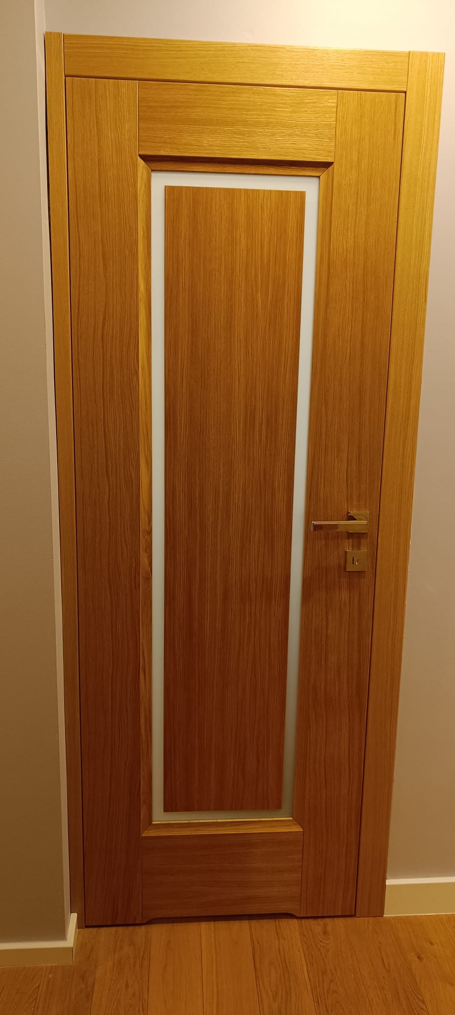 Dębowa, fornirowana ościeżnica z prostokątnym przeszkleniem i złotą klamką. Widoczna faktura drewna i podłoga w podobnym odcieniu. Minimalistyczny design.