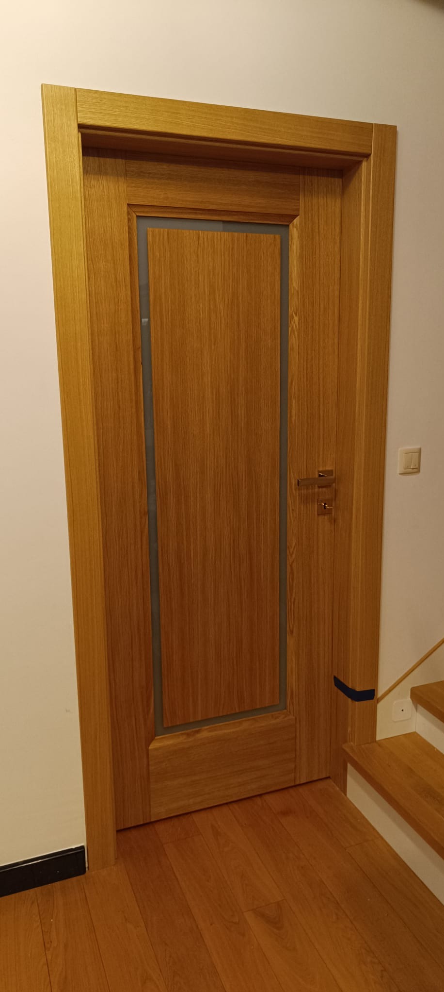 Dębowa, fornirowana ościeżnica z szarą intarsją i klamką na tle białej ściany i drewnianej podłogi. Widoczne schody. Ciepłe oświetlenie.