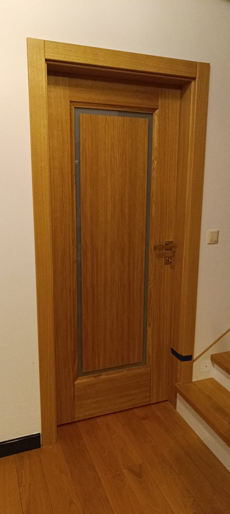 Drewniane drzwi wewnętrzne z przeszkleniem, widoczne na tle jasnej ściany i drewnianych schodów. Klamka w kolorze złotym. Ujęcie z bliska, prezentujące detale wykończenia.