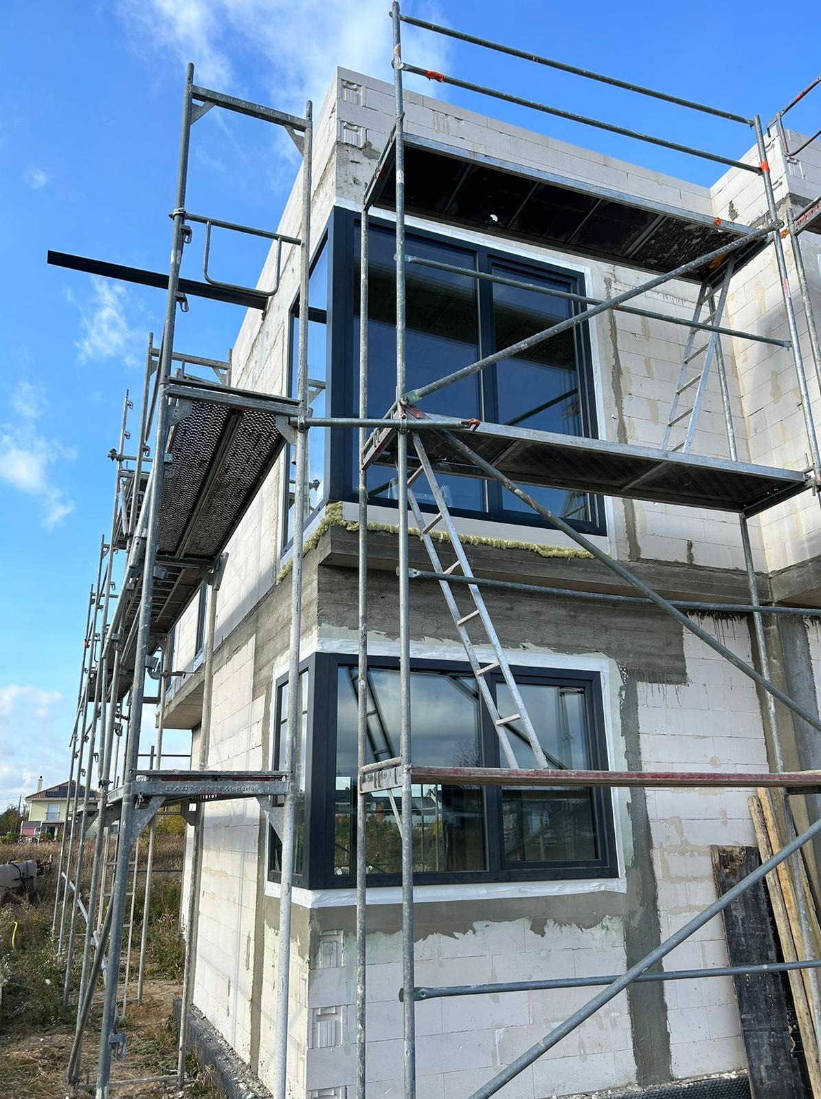 Montaż okien w surowym budynku z białego bloczka, widoczne rusztowania i drabina. Okna w ciemnych ramach, częściowo osadzone. Błękitne niebo w tle.