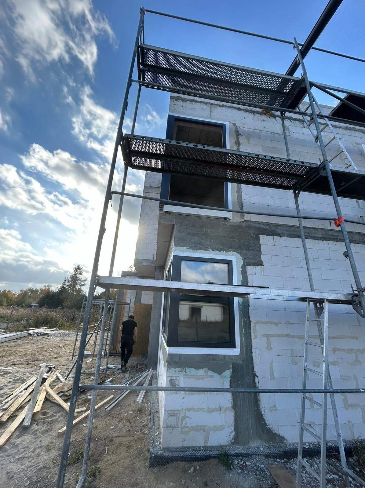 Montaż okien w surowym budynku z białego bloczka, widoczne rusztowania i drabina. Czarna rama okienna kontrastuje z jasnym murem. Osoba w tle. Słoneczny dzień.